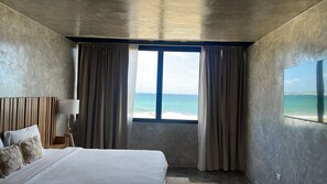 A bedroom with a bed, curtains, a nightstand, a lamp, and a view of the ocean.
