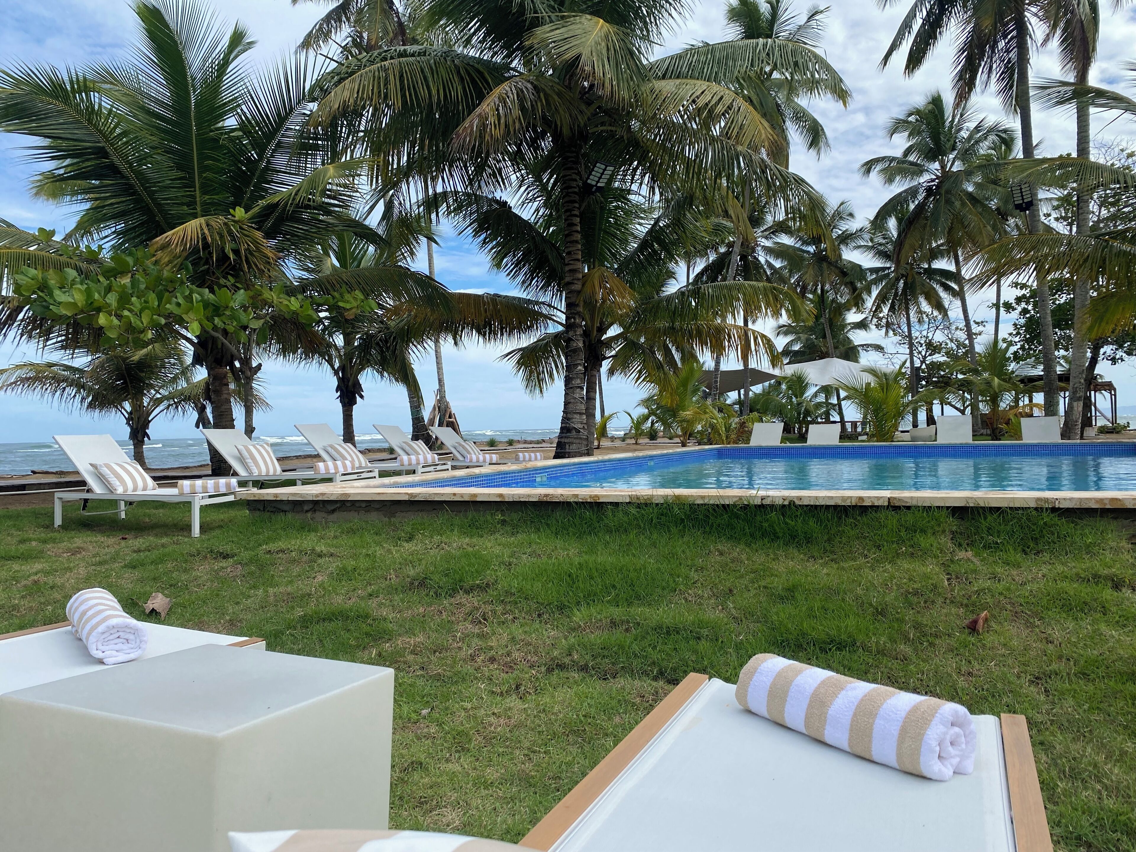 Zona junto a la piscina con sillones de descanso y una piscina, rodeada de palmeras y vista al océano.
