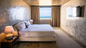 Ein Schlafzimmer mit Bett, Nachttisch mit Lampe, an der Wand montiertem Fernseher und einem Fenster mit Vorhängen.