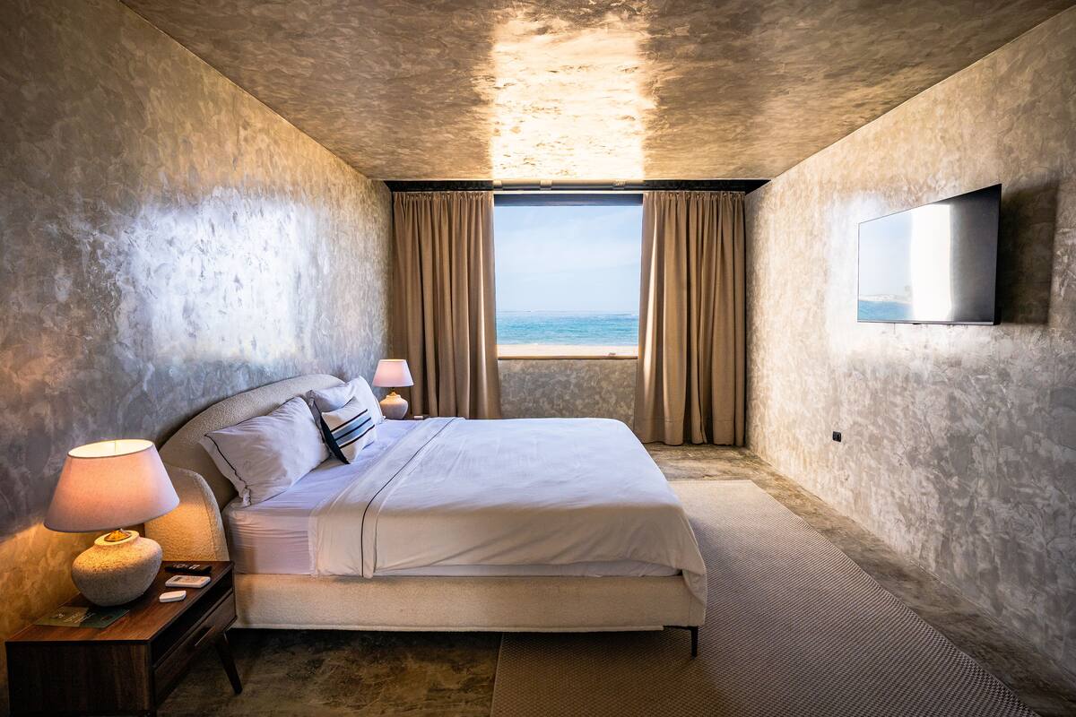 Un dormitorio con cama, una mesita de noche con lámpara, televisor montado en la pared y una ventana con cortinas.