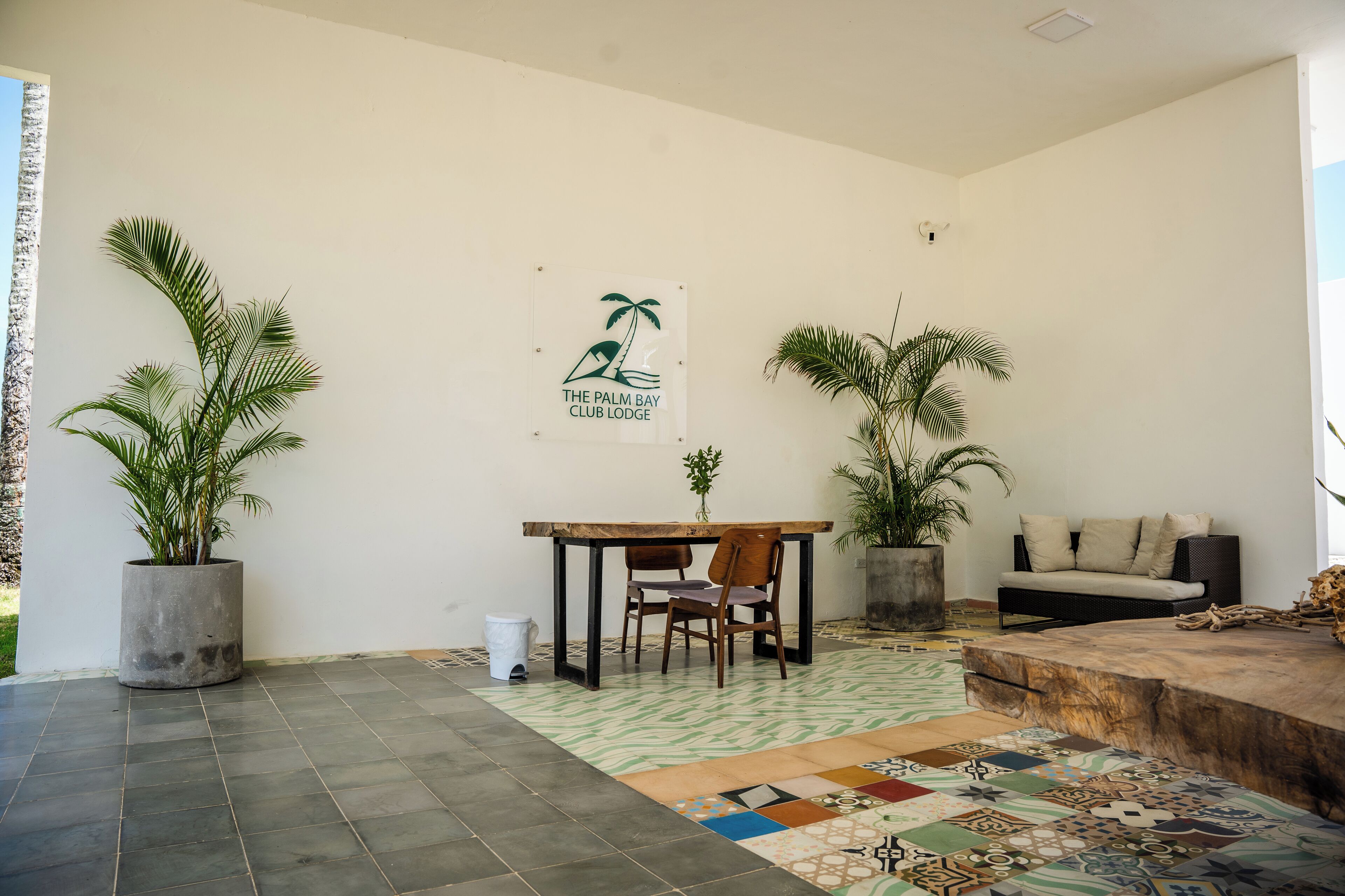 A modern living room with a wooden table, two chairs, a sofa, and two large potted plants. A sign with palm tree and waves design is on the wall.