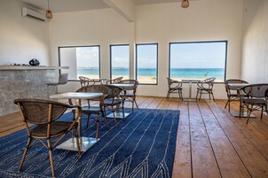 Uma área de refeições à beira-mar com cadeiras de vime, uma mesa e grandes janelas com vista para o oceano.
