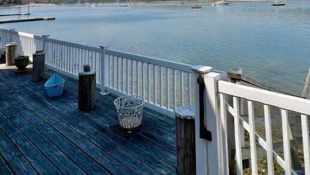 A wooden deck with a railing overlooking a calm body of water, boats in the distance, and a clear sky.