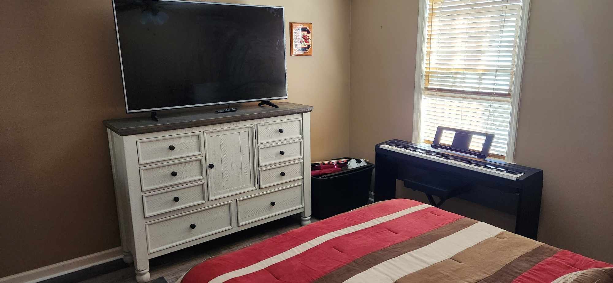 A bedroom with a television on a dresser, a bed with a striped blanket, a keyboard, and a window with blinds.