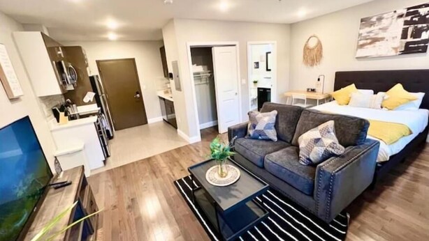 A modern living room with a sofa, coffee table, and a TV. There is a bed with pillows and a bedside table. The room has wooden flooring and a kitchen area in the background.