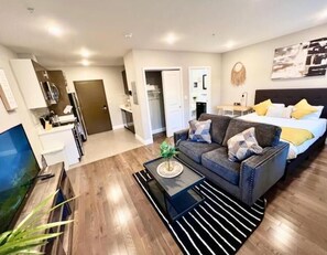 Ein modernes Wohnzimmer mit Sofa, Couchtisch und Fernseher. Ein Bett mit Kissen und ein Nachttisch sind ebenfalls vorhanden. Der Boden ist mit Holz ausgelegt, und im Hintergrund ist eine Küchenzeile zu sehen.