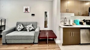 A modern living room with a grey sofa, a small red table, and a kitchen area with stainless steel appliances and wooden cabinets. - 306 Deluxe King suite in the center city (Philadelphia)