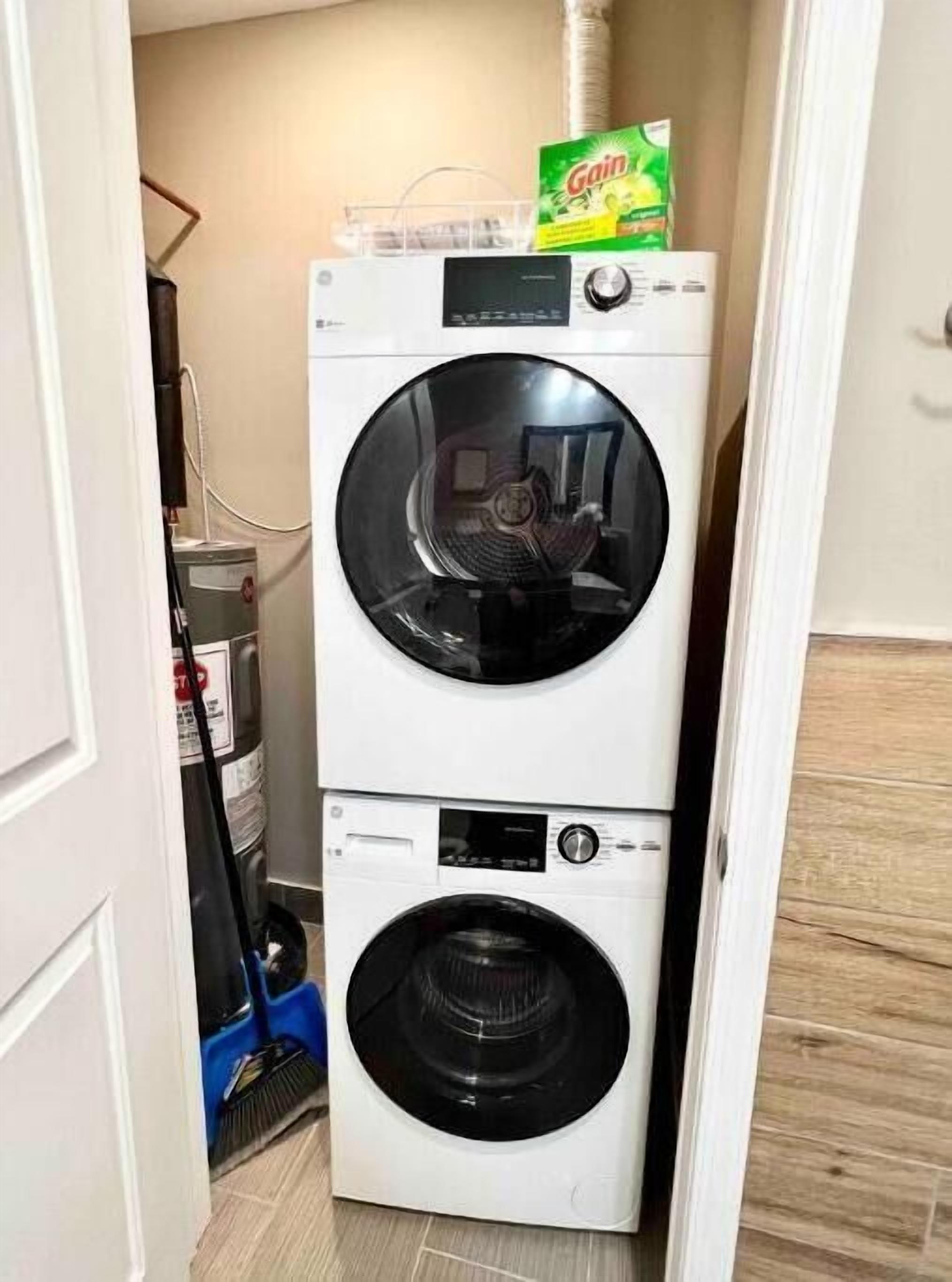 Eine kompakte, weiße, übereinander gestellte Waschmaschine und Trockner in einem schmalen Waschraum, mit einem sichtbaren Schlauch und einem Besen.