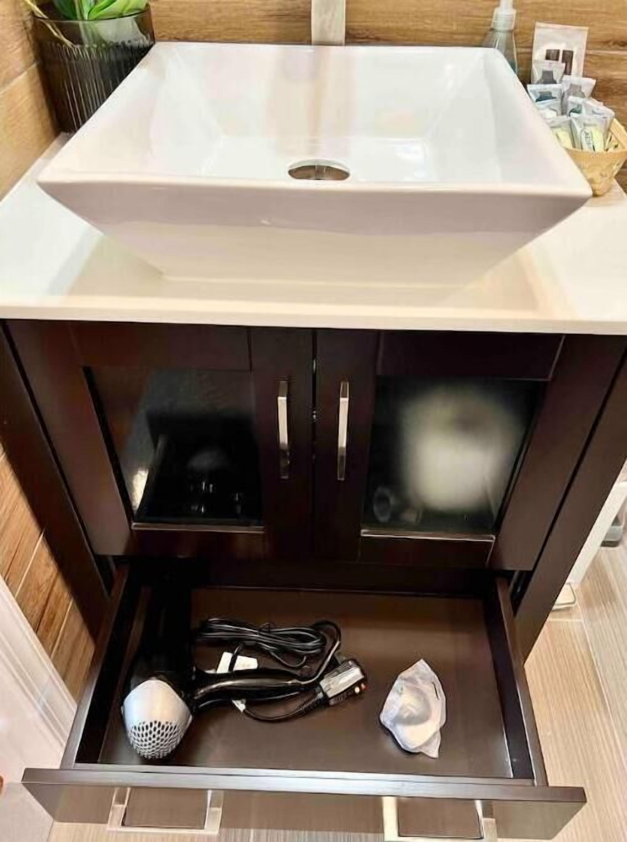 A bathroom vanity with a white sink, dark wooden cabinets, and a drawer containing a hairdryer, a power cord, and a tissue.