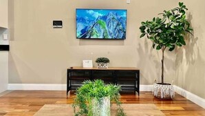 A living room with a wooden floor, a large flat-screen TV mounted on the wall, a console table with a plant, and a tall potted plant. - 402 Stylish luxe Apt at center city self check-in (Philadelphia)