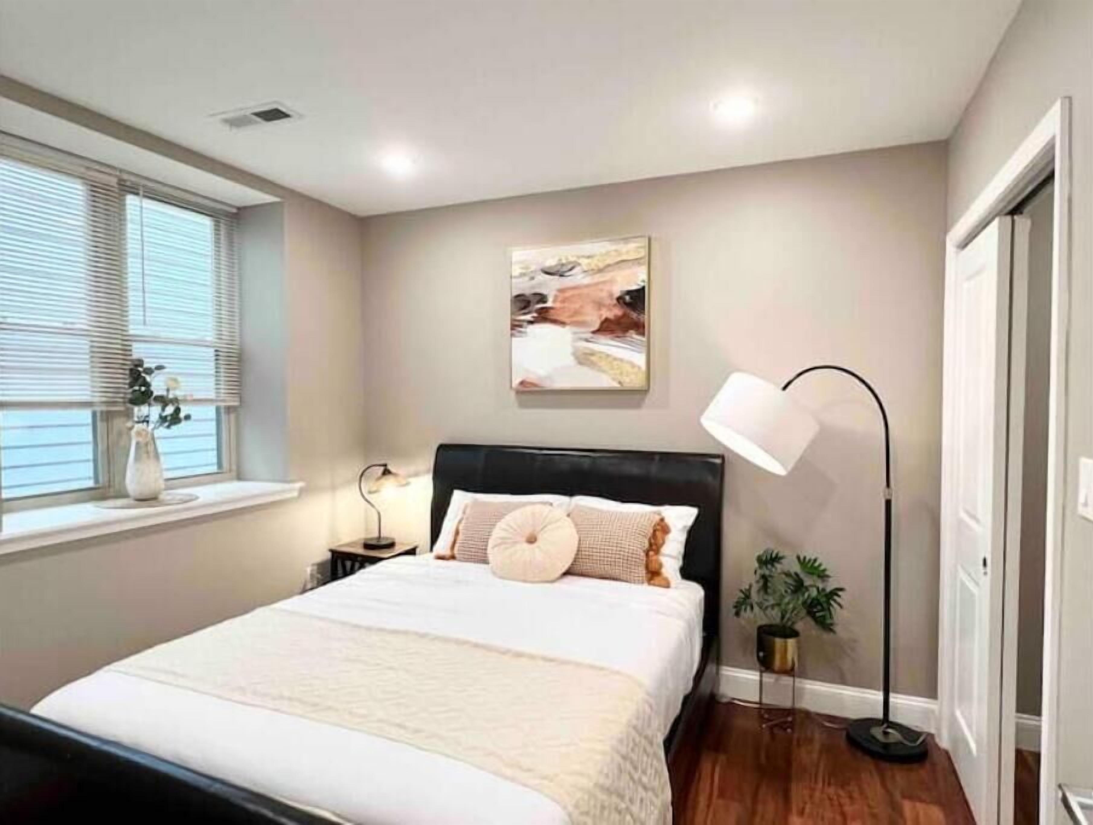 Un dormitorio con cama, mesitas de noche, lámpara de pie, ventana con persianas y un cuadro colgado en la pared.