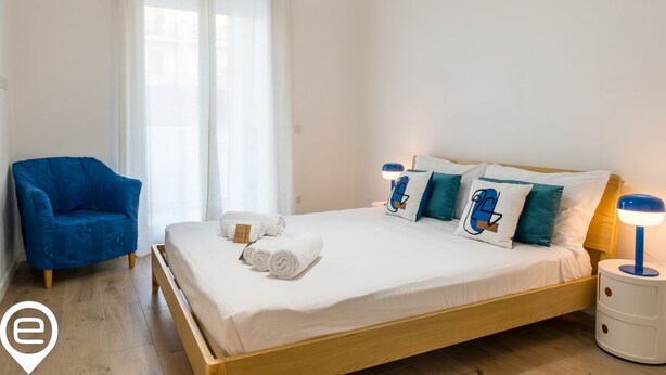 Un dormitorio con cama, un sillón azul, una mesita de noche con lámpara y una ventana con cortinas.