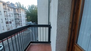 Un balcón con barandilla negra, suelo de tejas rojas y vista a una calle con coches aparcados y edificios al fondo.