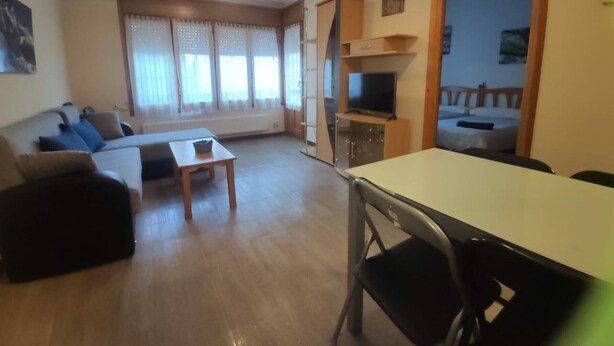 Una sala de estar con sofá, una mesa y un televisor. Hay una ventana con cortinas y una puerta que da a otra habitación.