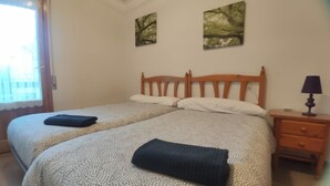 Cama doble con cabecera de madera, dos almohadas y una manta doblada a cada lado. Mesita de noche con lámpara y cajón. Dos cuadros adornan la pared sobre la cama.