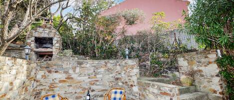 Une terrasse extérieure avec des murs en pierre, une cheminée en pierre et deux fauteuils en osier, une bouteille de vin étant posée sur une table.