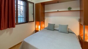Un dormitorio con una cama, dos almohadas, una mesita de noche, una ventana con cortinas y una estantería con una planta.