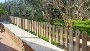Un sendero de concreto largo y estrecho bordeado por una cerca de madera, que conduce a una zona con césped y árboles.