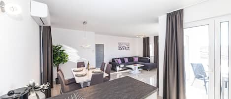 Een moderne woonkamer met een eethoek, een grijze bank en een groot raam met gordijnen.