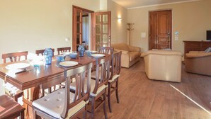 Zona pranzo con tavolo in legno apparecchiato per quattro, circondato da sedie imbottite. C'è un divano e un armadio nella zona soggiorno.
