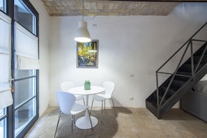 A room with a round table, four white chairs, a hanging light fixture, a framed picture on the wall, and a staircase.