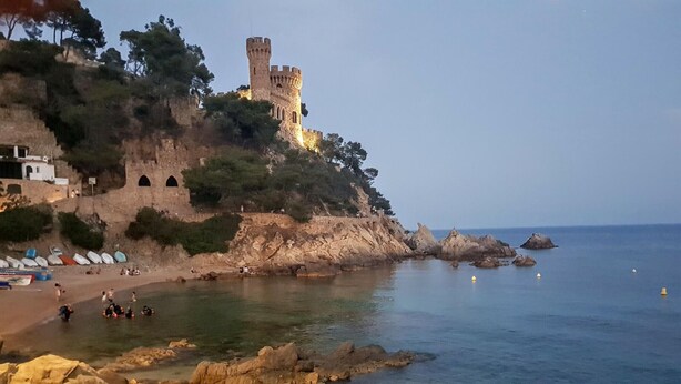 A coastal castle with a tower, rocky shoreline, clear water, and beach with umbrellas. - Beachfront Apartment with Wifi and Easy Access to Attractions (Lloret de Mar)