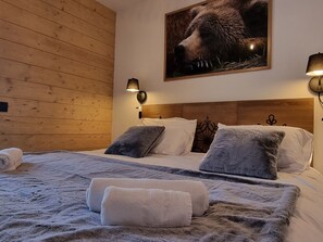 Habitación de hotel con paredes de paneles de madera, una cama grande con ropa de cama gris y blanca, dos lámparas fijadas a la pared y un cuadro de un oso.
