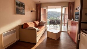 Ein kompakter Wohnbereich mit Sofa, kleinem Tisch, Küchenzeile und einer Schiebeglastür, die auf eine Außenpatio führt.