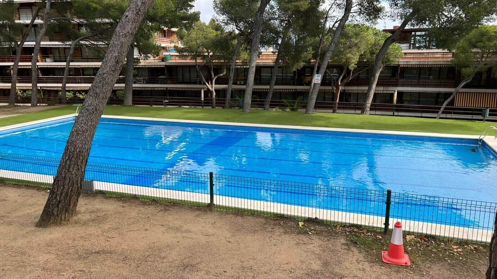 A rectangular swimming pool with a surrounding fence, trees, and a building in the background.