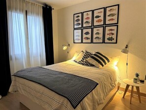 A neatly made bed with a striped blanket, a wall with framed fish pictures, a bedside lamp, and a small table with a plant.