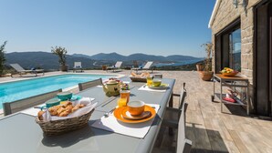 A poolside breakfast setup with a table, chairs, and a view of mountains.