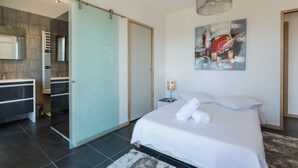 A modern bedroom with a large bed, a bathroom with a glass shower, and a painting on the wall.