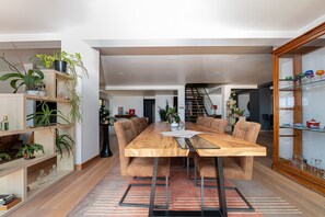A modern dining area with a wooden table, cushioned chairs, and a glass-top table. There are shelves with plants and a glass cabinet with decorative items.
