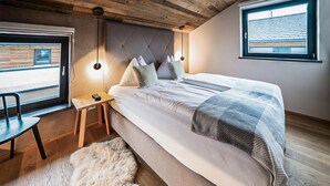 Ein Schlafzimmer mit Bett, einem Sessel, einem kleinen Tisch, einem Fellteppich und einem Fenster mit Jalousien.
