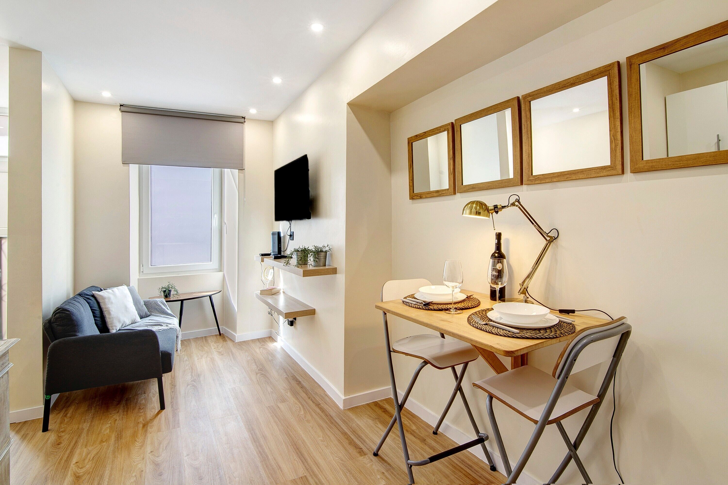 A modern living room with a sofa, a dining table with chairs, a TV mounted on the wall, and a window with a blind.