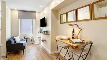 A modern living room with a sofa, a dining table with chairs, a TV mounted on the wall, and a window with a blind.