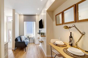 A modern living room with a sofa, a dining table set for two, a TV mounted on the wall, and a window with a view.