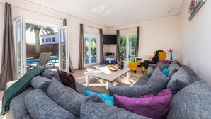A living room with a grey sofa, a coffee table, and a TV. There are curtains, a chair, and a small table with a plant. - Villa 'Amarador' with Private Pool, Wi-Fi and Air Conditioning (Santanyí)