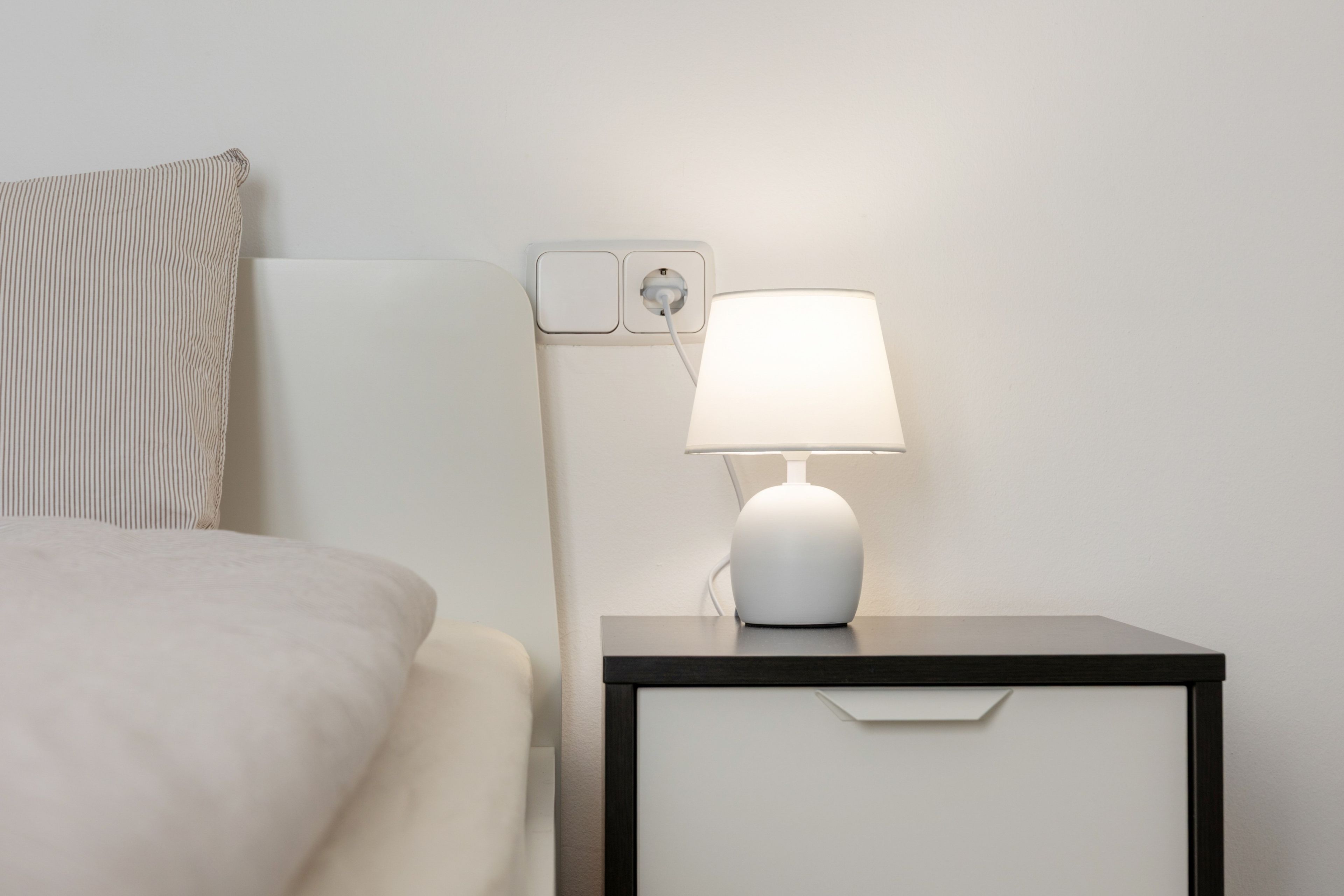 Una cama bien hecha con una colcha blanca y un almohadón a rayas. Junto a la cama, una mesita de noche con una lámpara blanca y un enchufe.