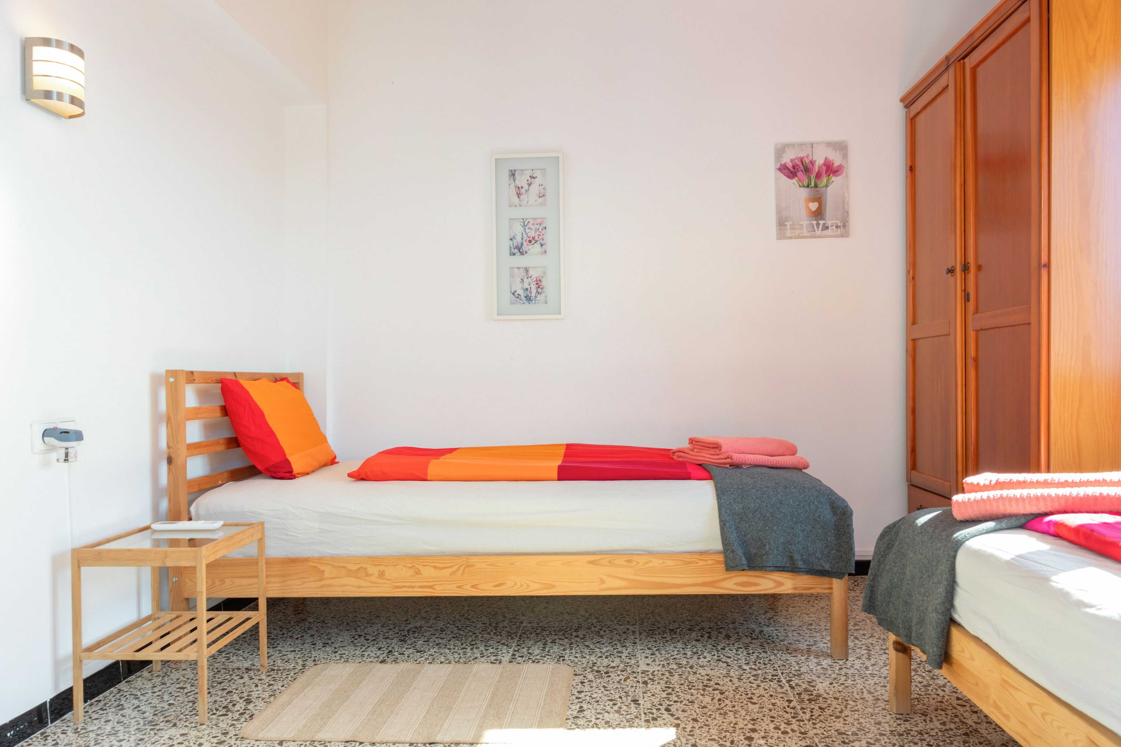 Un dormitorio con una cama de madera, una mesita de noche pequeña, una lámpara de pared, una obra de arte floral enmarco y un armario.