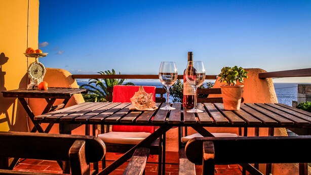 Une table en bois avec deux verres à vin et une bouteille, une plante en pot et une balance, le tout sur un balcon donnant sur la mer.