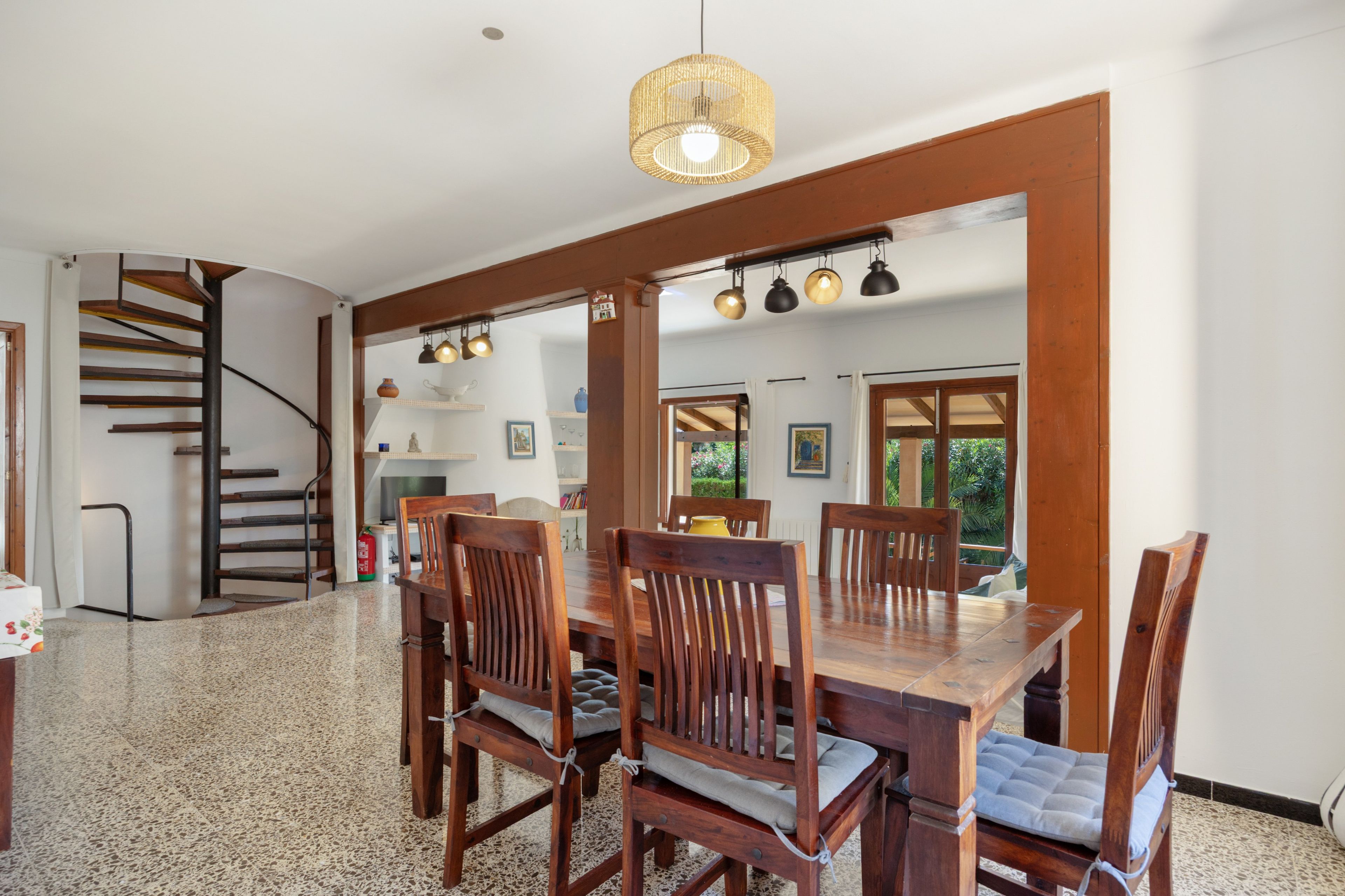Un área de comedor con una mesa y sillas de madera, una escalera y una cocina visible a través de un arco.