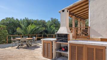Eine Außenpatio mit Steinboden, einem Grill, Holzmöbeln und Blick auf üppiges Grün.