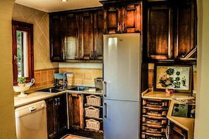 A kitchen with wooden cabinets, a white refrigerator, a dishwasher, and a window with a view of greenery. - Holiday Home 'Villa Buena Vista' with Sea View, Wi-Fi and Air Conditioning (Cala Pi)