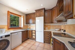 Une cuisine avec des meubles en bois, un réfrigérateur blanc, un lave-vaisselle et une fenêtre donnant sur un paysage verdoyant.