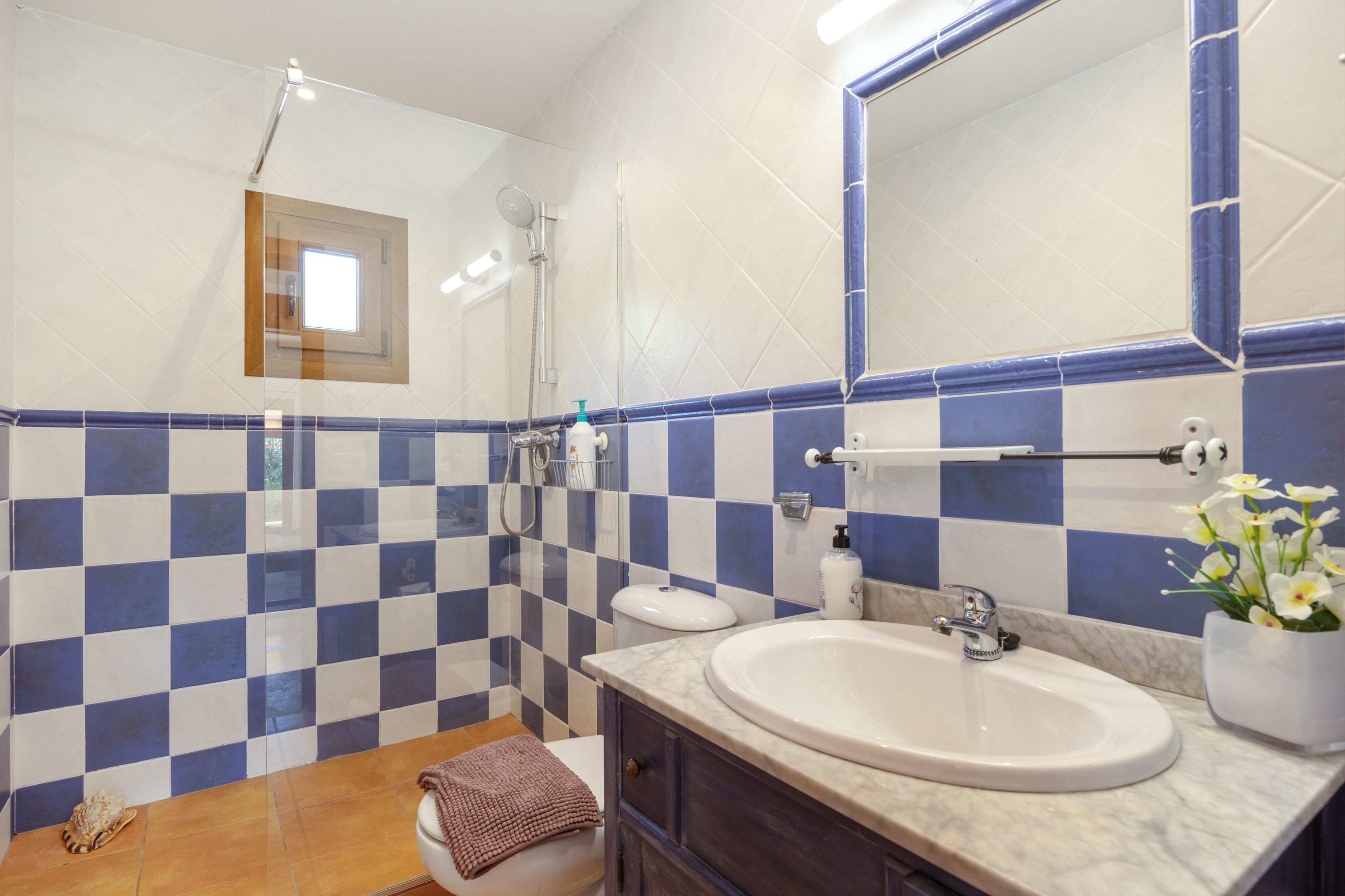 Un baño con pared de baldosas a cuadros, inodoro, lavamanos con encimera de mármol, espejo y ducha.