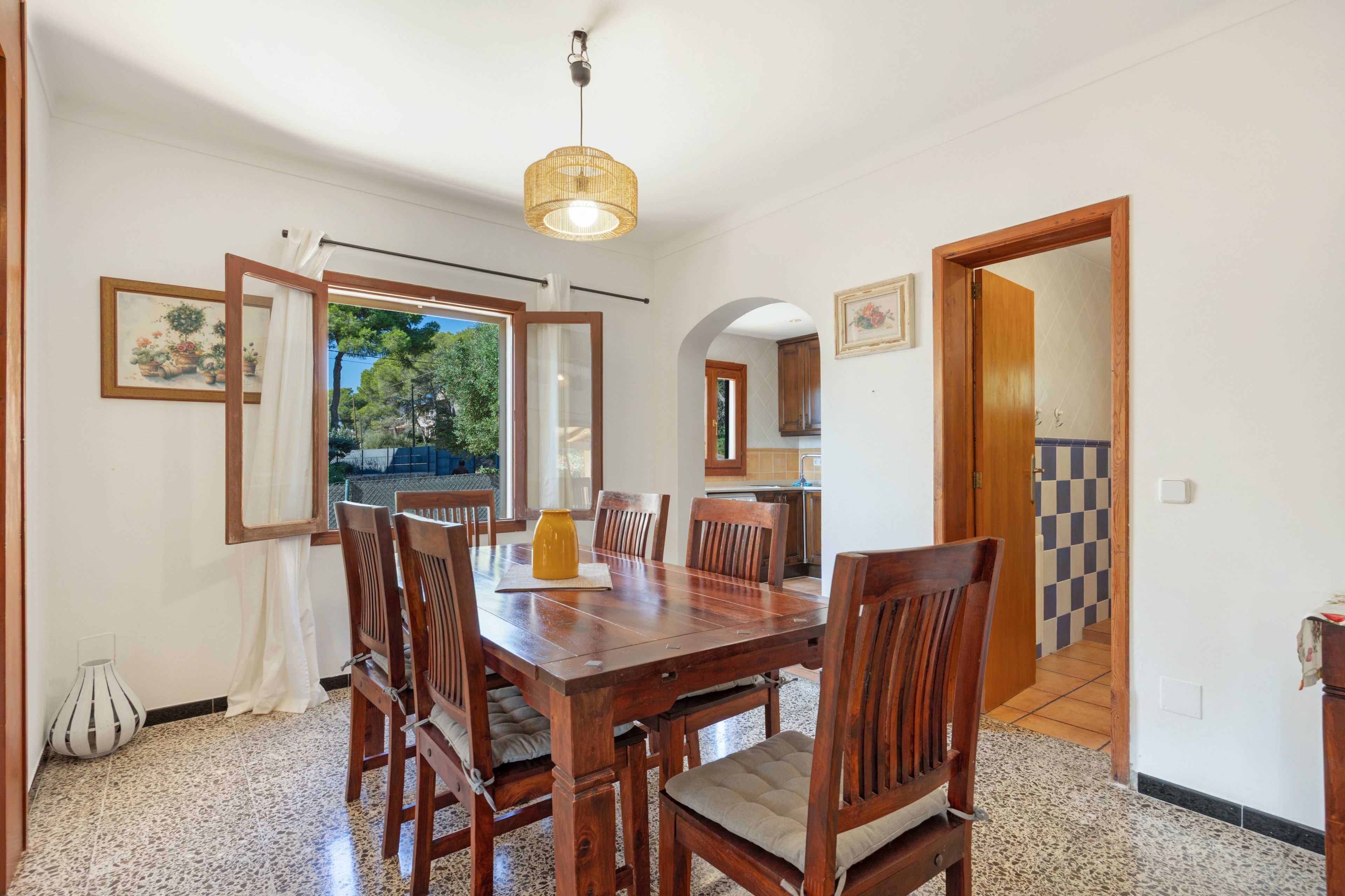 Un comedor con mesa y sillas de madera, una lámpara colgante y vistas a través de un ventanal abierto.