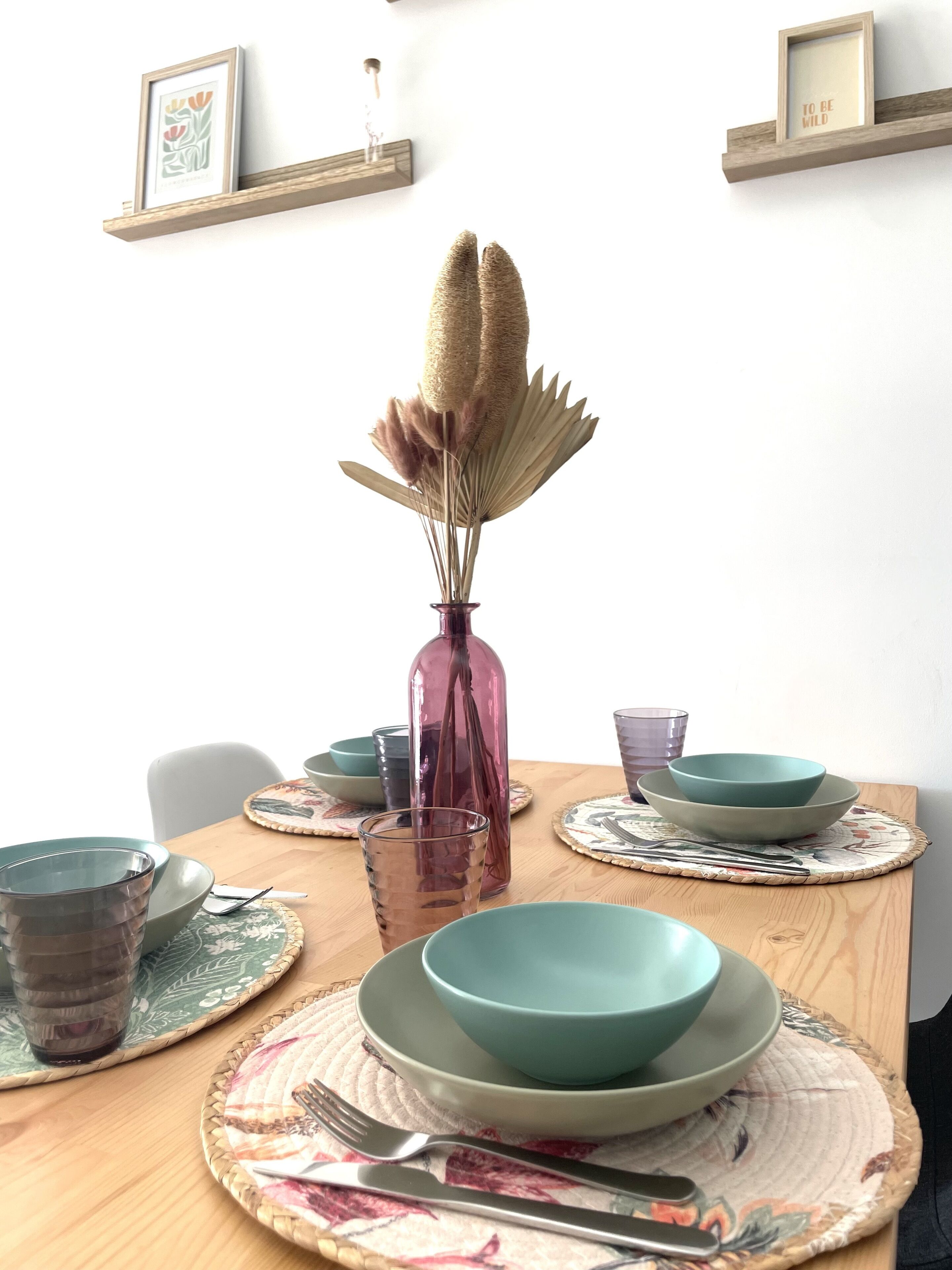 Une table à manger dressée avec des bols, des assiettes et des couverts de couleur turquoise, un vase rose contenant des fleurs séchées et un verre d’eau.