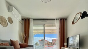 Una sala de estar moderna con un sofá gris, una mesa de centro, un televisor sobre un mueble blanco y un ventanal con cortinas.