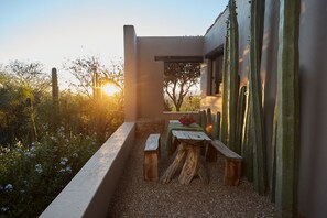 A patio with a wooden table and benches, surrounded by cacti and flowering plants, with a view of trees and the sun setting in the background.